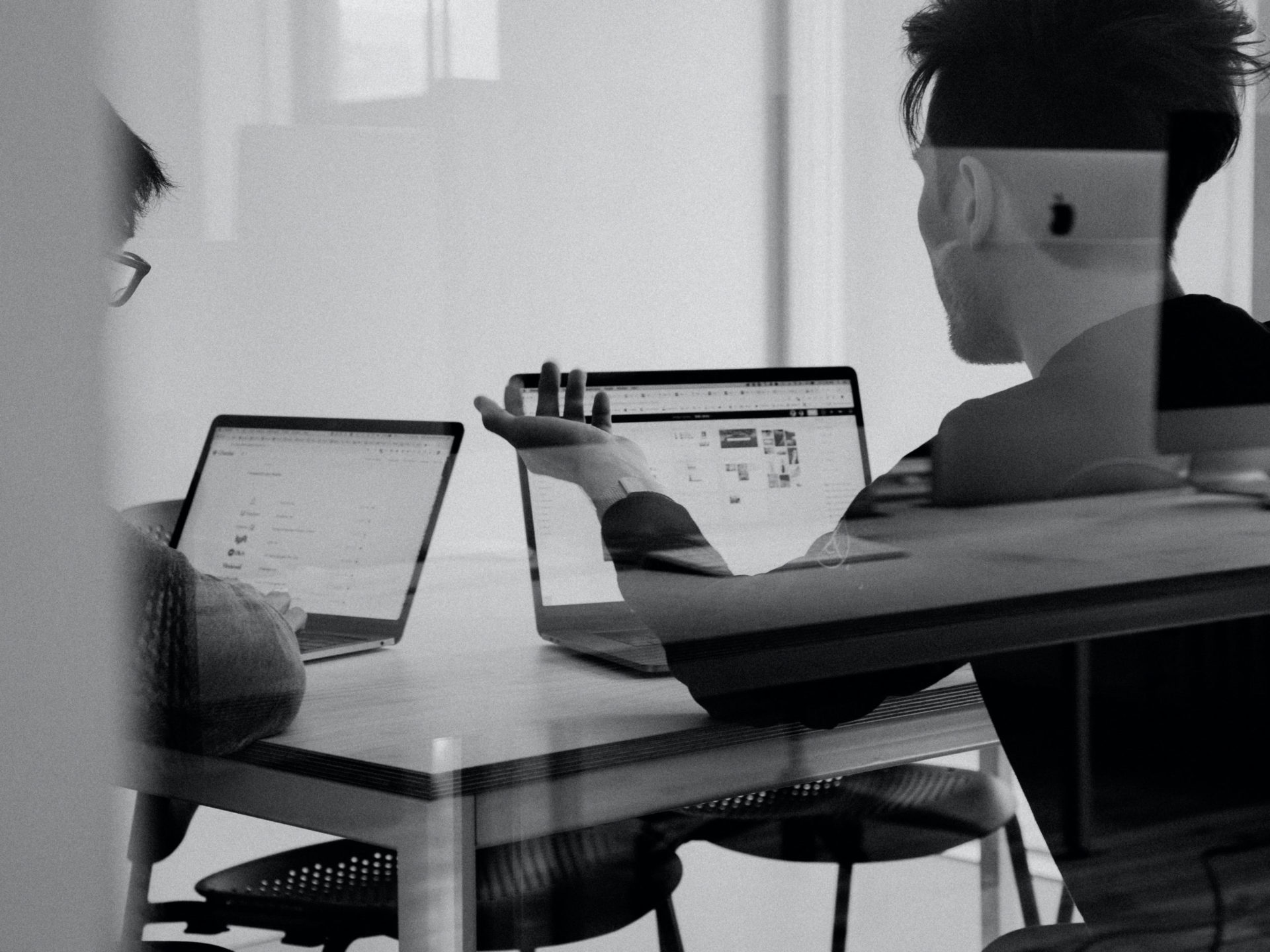 Two black and white figures sit at a table discussing things while using their laptops. There's reflection in the foreground that indicate the viewer is outside the room